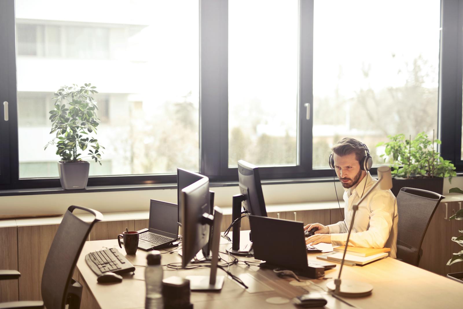 Un homme d'affaires est assis à un bureau, utilisant plusieurs ordinateurs et un casque, dans un bureau moderne bien éclairé.